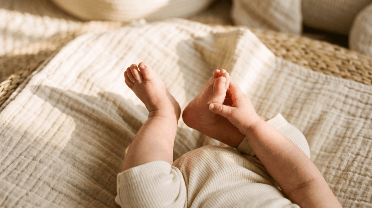 Bébé attrape ses pieds allongé sur le dos sur un tapis d'éveil, lumière rasante sur un lange en coton – Treelys®