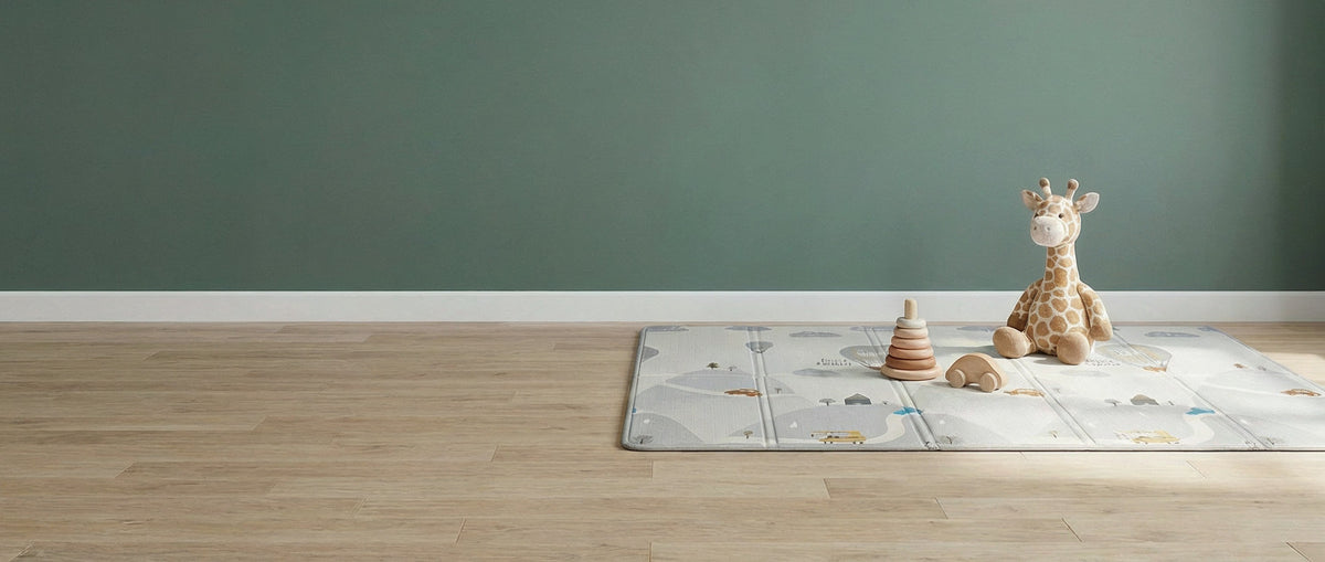Peluche girafe assise sur un tapis d'éveil Treelys déplié devant un mur vert sauge